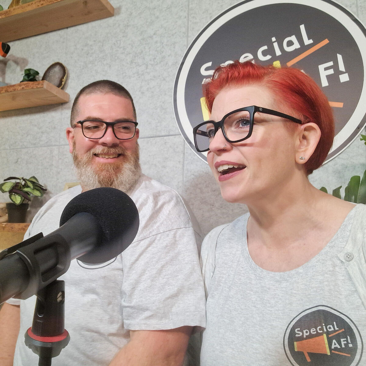 An image of a bearded man smiling and a red-haired woman talking into a microphoneat a podcast desk